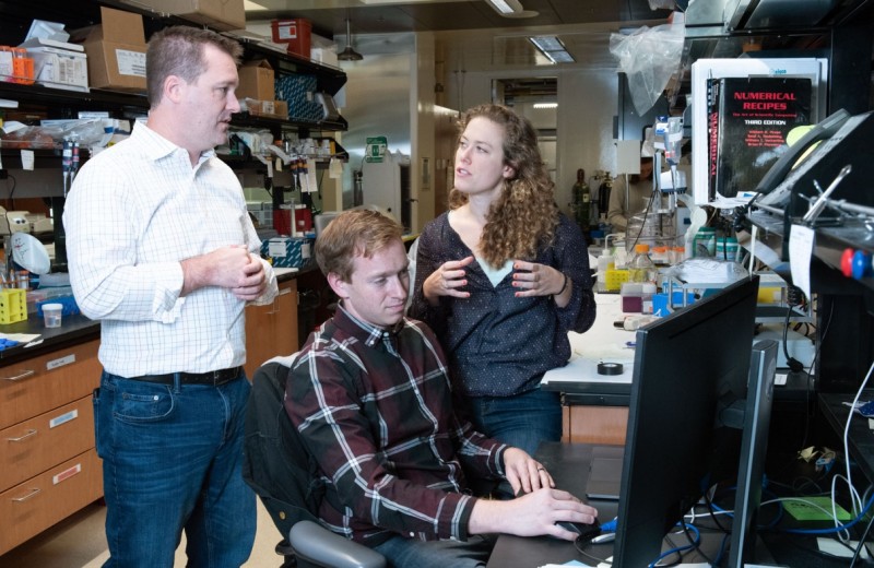 Todd McDevitt and Ashley Libby standing behind David Joy who is working on a computer in the lab